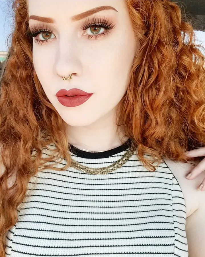 Femme aux cheveux roux bouclés, maquillage soigné, regard intense, portant un haut rayé noir et blanc, assise dans une voiture.