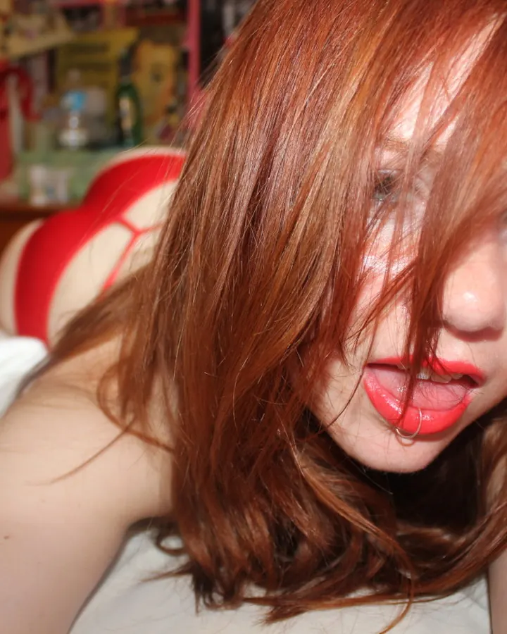 Femme aux cheveux roux, en haut rouge, allongée sur un lit blanc, cheveux devant le visage, regardant l'appareil, lèvres rouges.