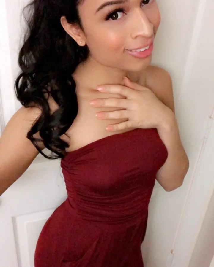 Femme aux cheveux noirs ondulés, souriante, portant une robe rouge bordeaux sans bretelles, main sur la poitrine, devant une porte blanche.