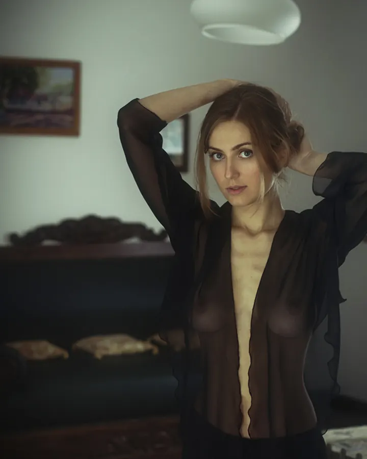 Femme aux cheveux bruns, en chemise noire transparente, main dans les cheveux, regardant l'appareil, dans une pièce sombre avec un canapé.