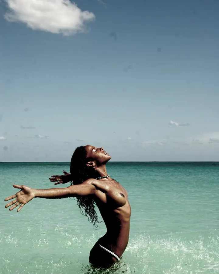 Femme noire aux cheveux longs dans l'eau turquoise, bras ouverts vers le ciel, corps mouillé sous le soleil.
