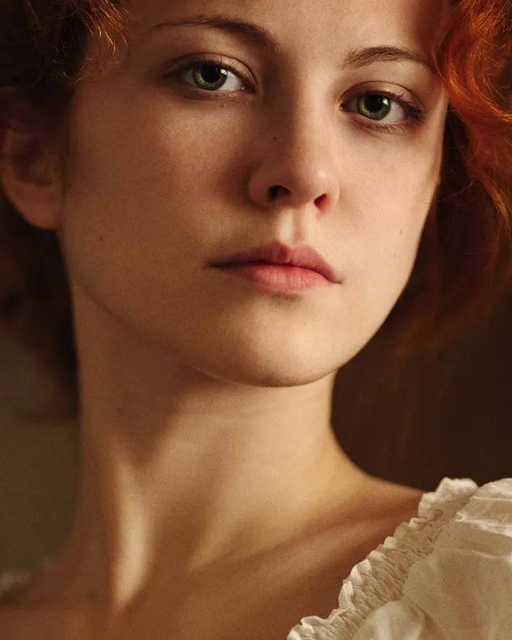Femme rousse aux yeux gris, portant un haut noir, regardant directement l'objectif avec une expression neutre, sur fond clair.