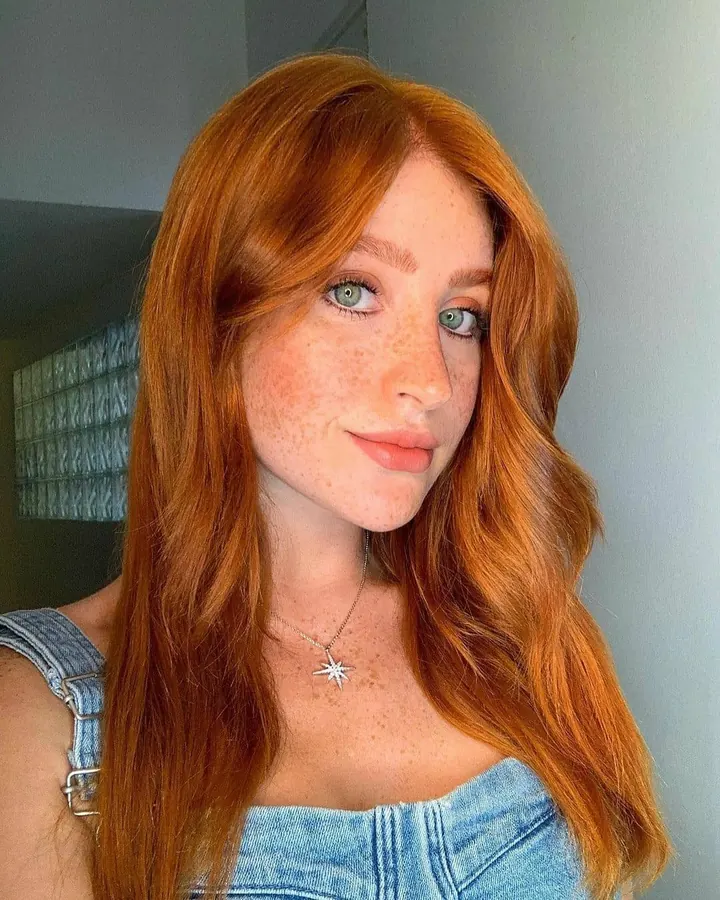 Femme rousse aux yeux gris, portant un haut noir, regardant directement l'objectif avec une expression neutre, sur fond clair.