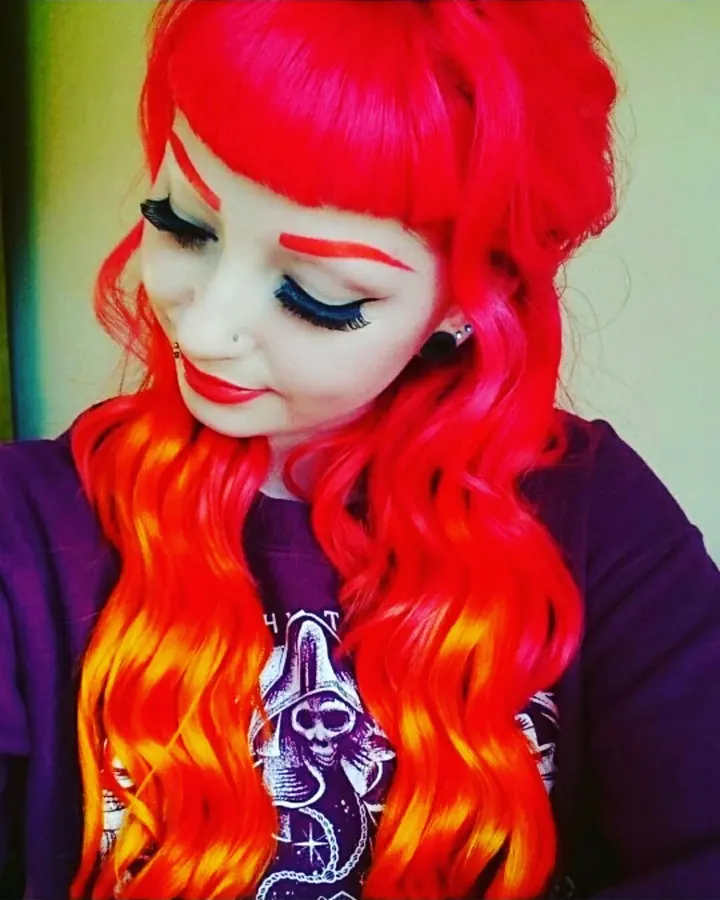 Femme aux cheveux rouges flamboyants, maquillage exagéré avec sourcils rouges, lèvres rouges, nez percé, vêtue d'un t-shirt violet à motif squelette.