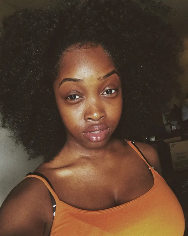 Femme africaine aux cheveux afro volumineux, portant un haut orange décolleté, regardant directement l'objectif avec une expression assurée.