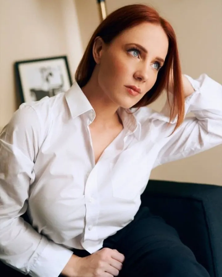 Femme en chemise blanche, assise, main sur la nuque, regardant l’appareil.