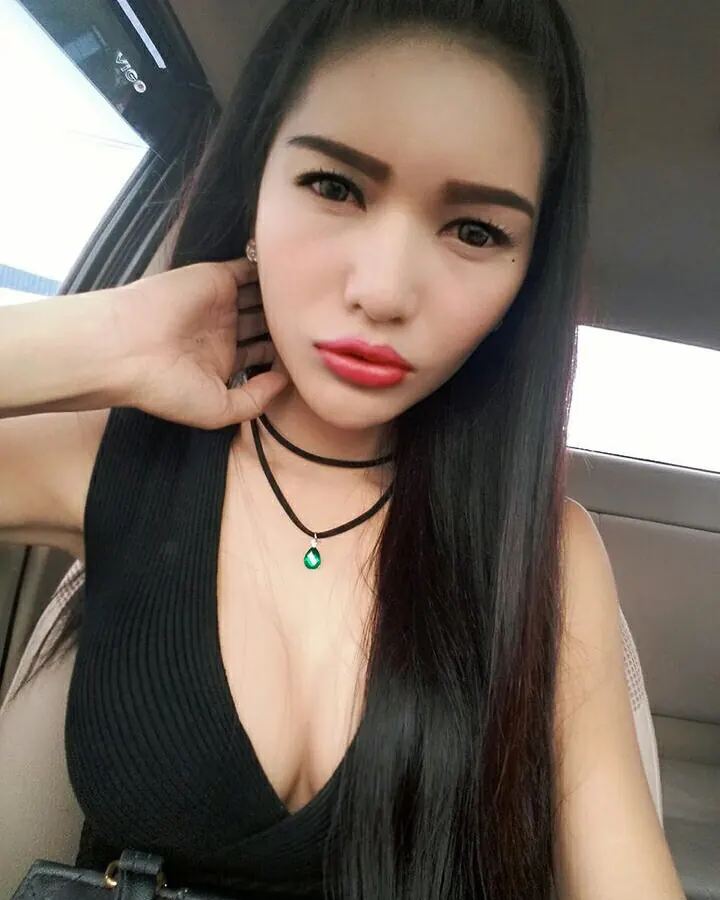 Femme en tenue noire, collier vert, assise dans une voiture, main sur la joue.