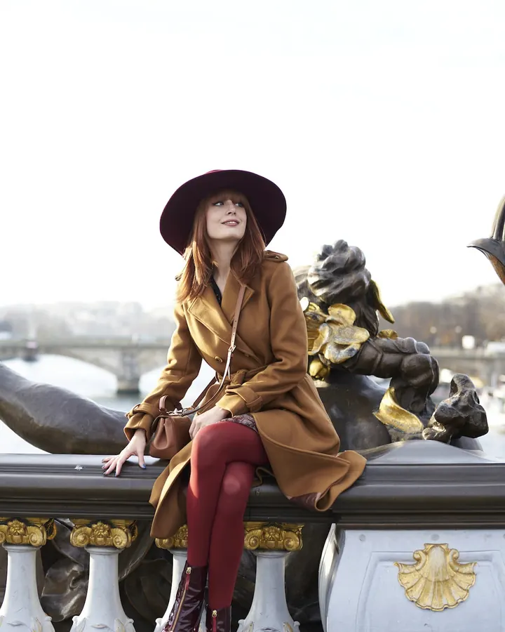 Femme blonde en robe marron, assise sur un pont décoratif avec un chapeau violet, regardant vers le haut.