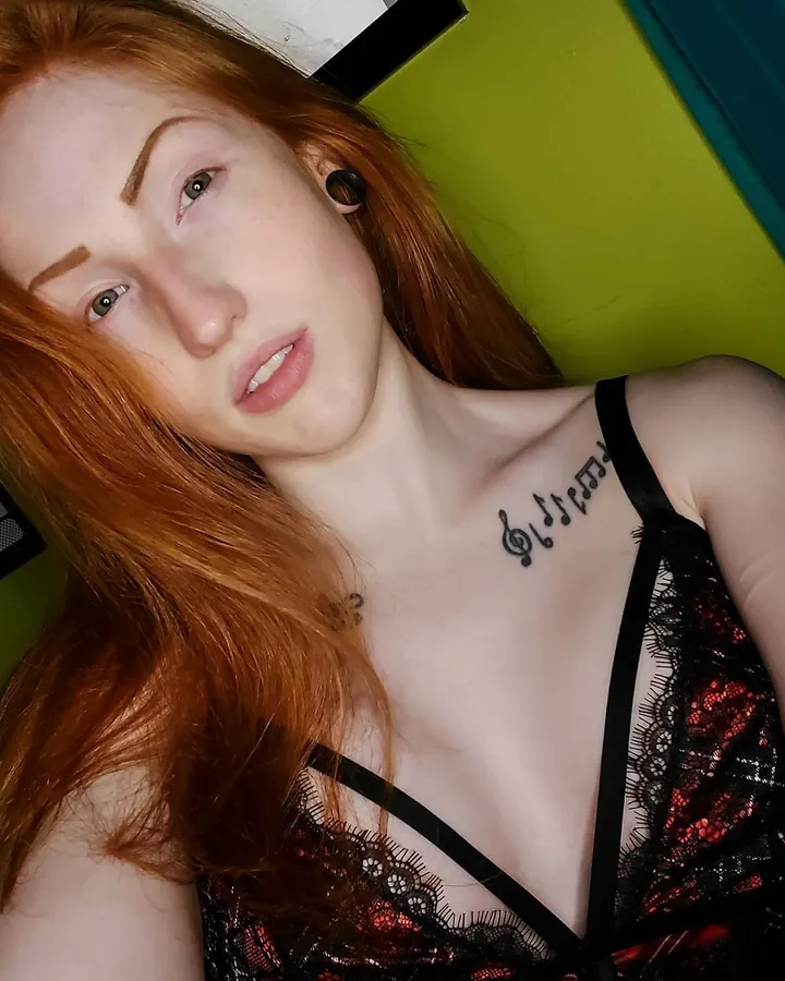 Femme aux cheveux roux, vêtue d’un haut noir à dentelle, tatouage musical sur la clavicule, regardant vers le haut