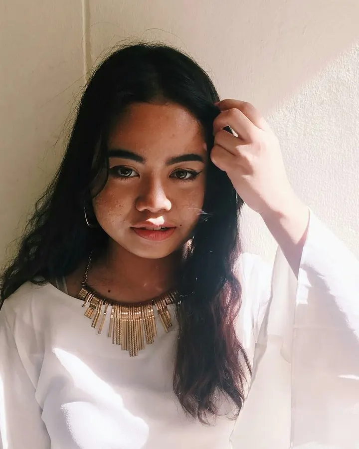 Femme latine en lingerie rouge, posant de profil contre un mur noir orné, les mains sur le mur.