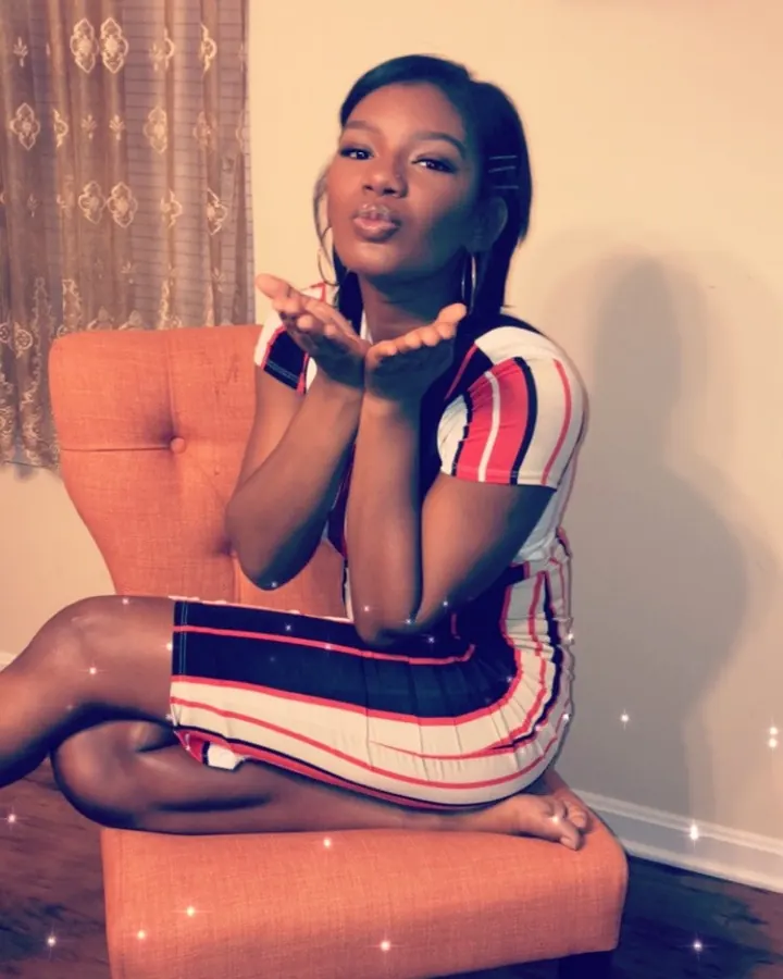 Femme aux cheveux noirs, assise sur une chaise orange, portant une robe rayée, faisant un geste de baiser avec les mains.