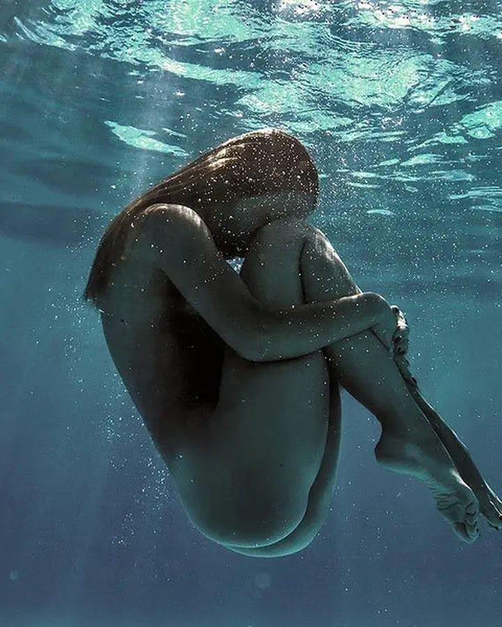 Femme nue sous l'eau, accroupie, les bras autour des genoux, avec des rayons de lumière filtrant depuis la surface.