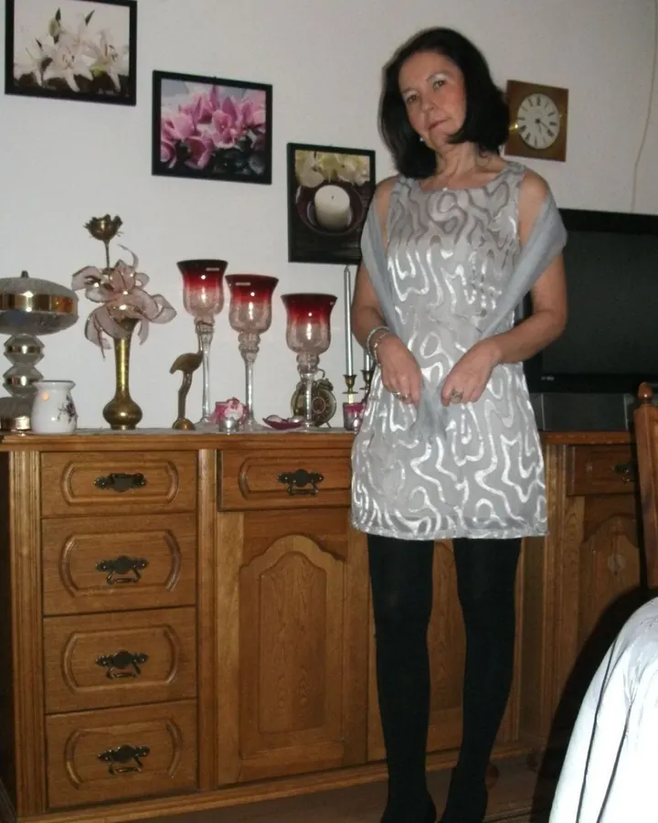 Femme aux cheveux noirs, debout dans une chambre, portant une robe grise à motifs et des collants noirs, devant une commode en bois.