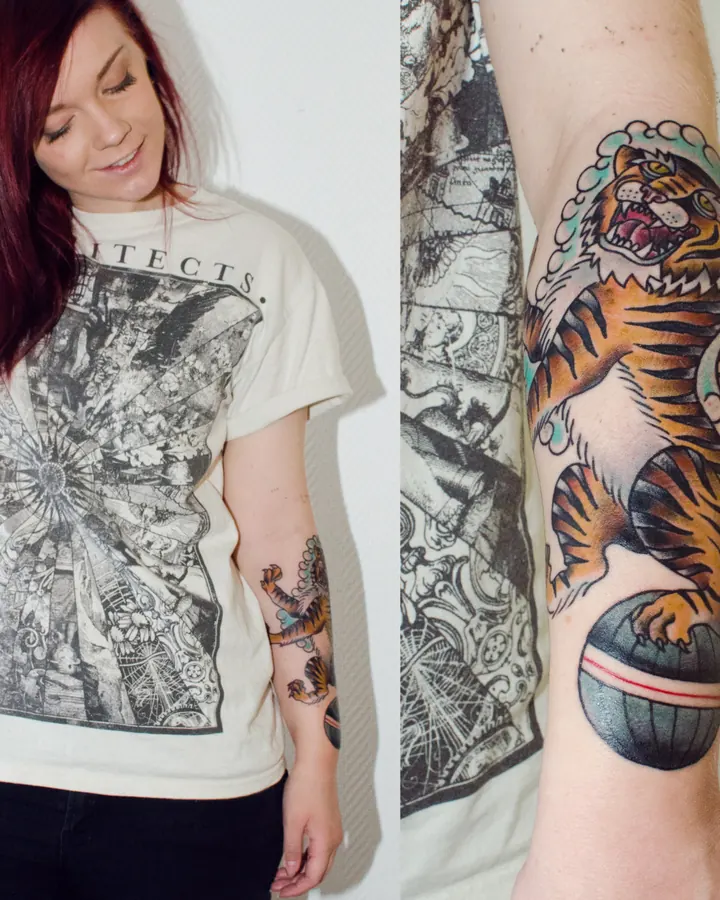 Femme aux cheveux roux, portant un t-shirt blanc avec un motif graphique, montrant un tatouage de tigre sur son avant-bras.