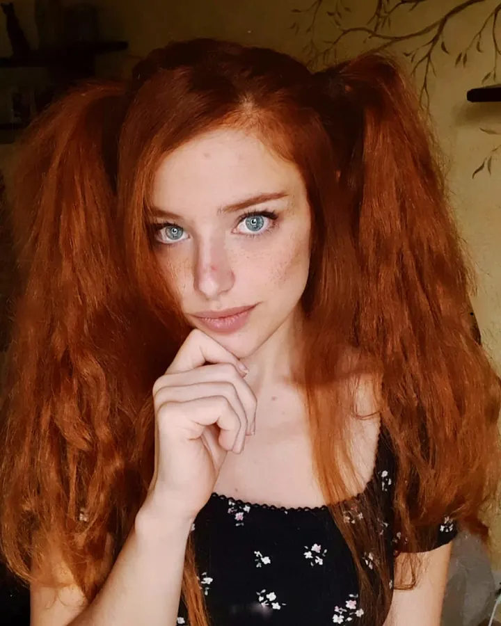 Femme aux cheveux roux, en robe noire, assise sur une couverture dans un champ, lisant un livre, main sur l'oreille.