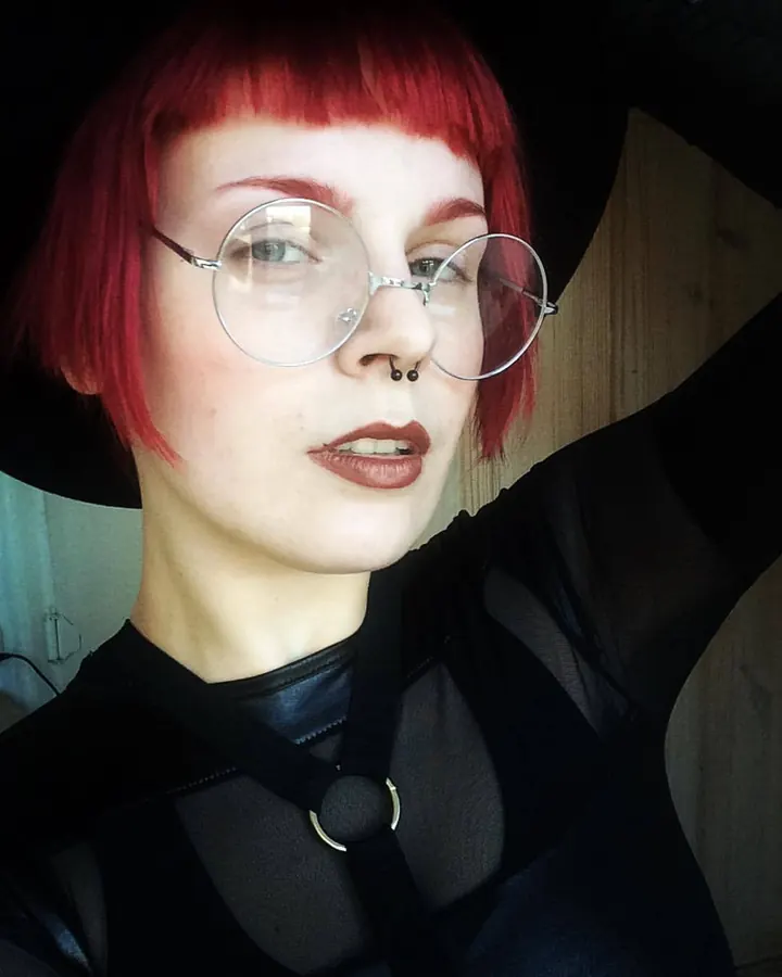 Femme aux cheveux roux, en robe rose, regardant l'objectif, main sur la joue, expression sérieuse.