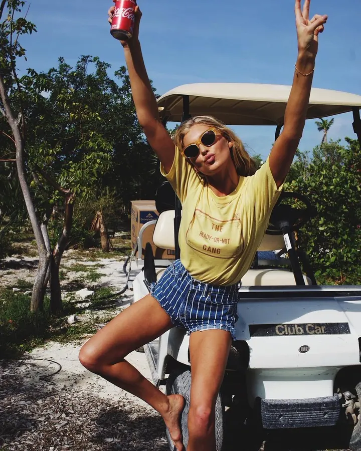 Femme blonde en tenue décontractée, assise sur un chariot de golf, tenant une canette de Coca-Cola, les bras levés, en plein air.
