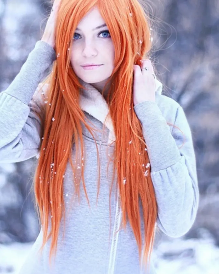 Femme aux cheveux roux, vêtue d'un pull gris, debout dans la neige, les mains dans les cheveux, regardant vers l'avant.
