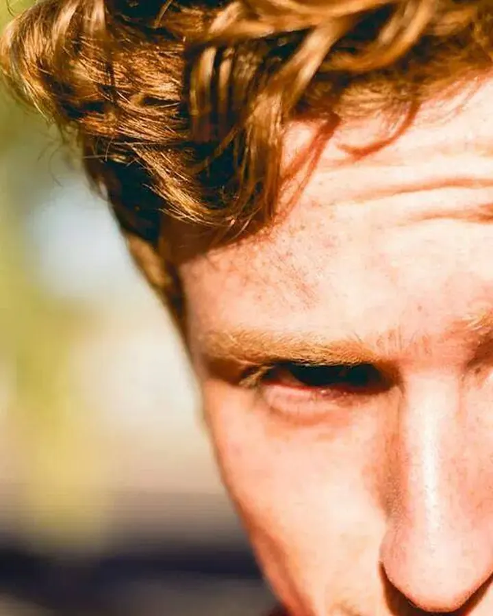 Visage d'un homme aux cheveux roux bouclés, regard intense, en gros plan sous la lumière du soleil.