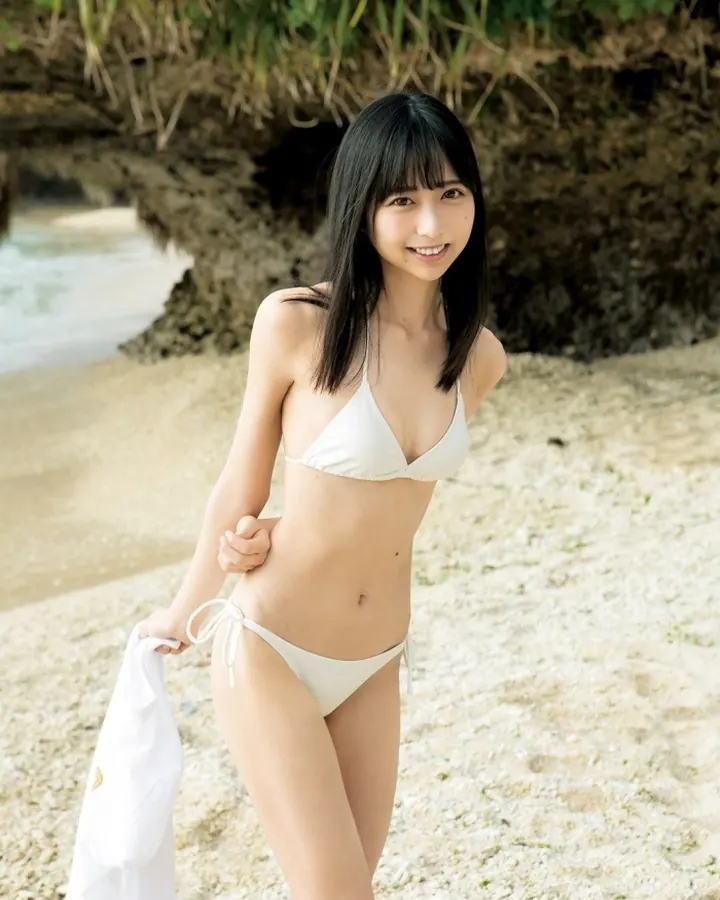 Femme asiatique en maillot blanc, souriante, tenant une serviette blanche sur une plage sablonneuse.