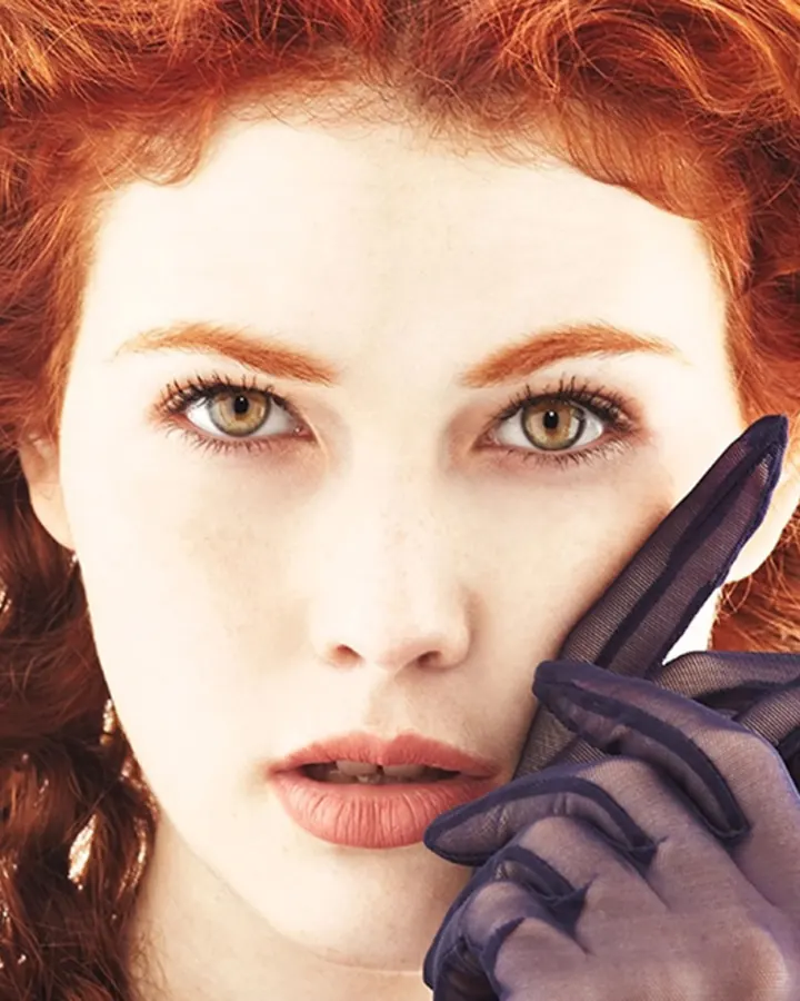 Femme aux cheveux noirs tressés, portant un haut orange et un jean bleu, tenant un sac marron, debout devant un mur avec des plantes.