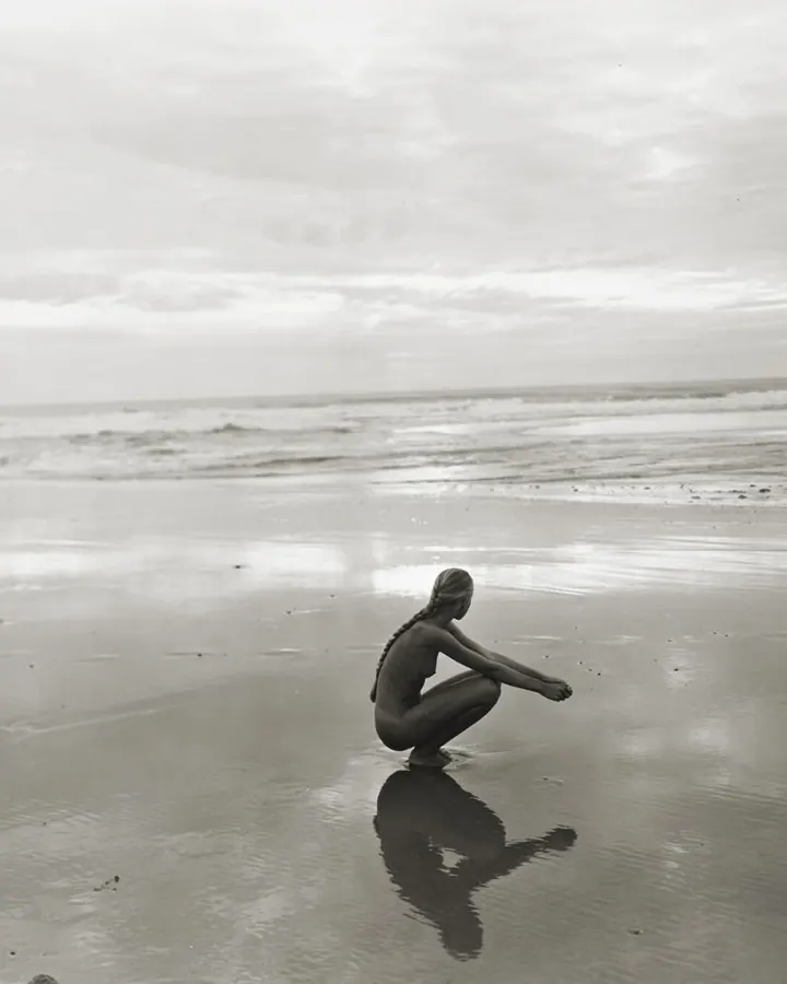 Silhouette féminine nue, agenouillée sur une plage mouillée, reflet dans l'eau, photo en noir et blanc.