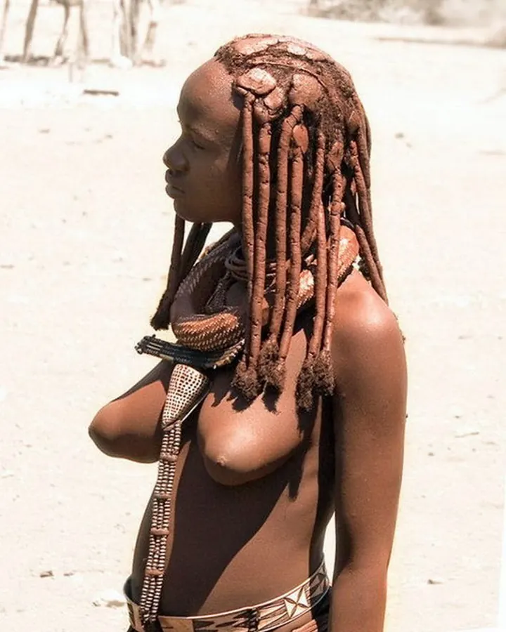 Femme africaine aux cheveux longs et tressés, peau foncée, torse nu, portant un collier et un bracelet, debout en plein air
