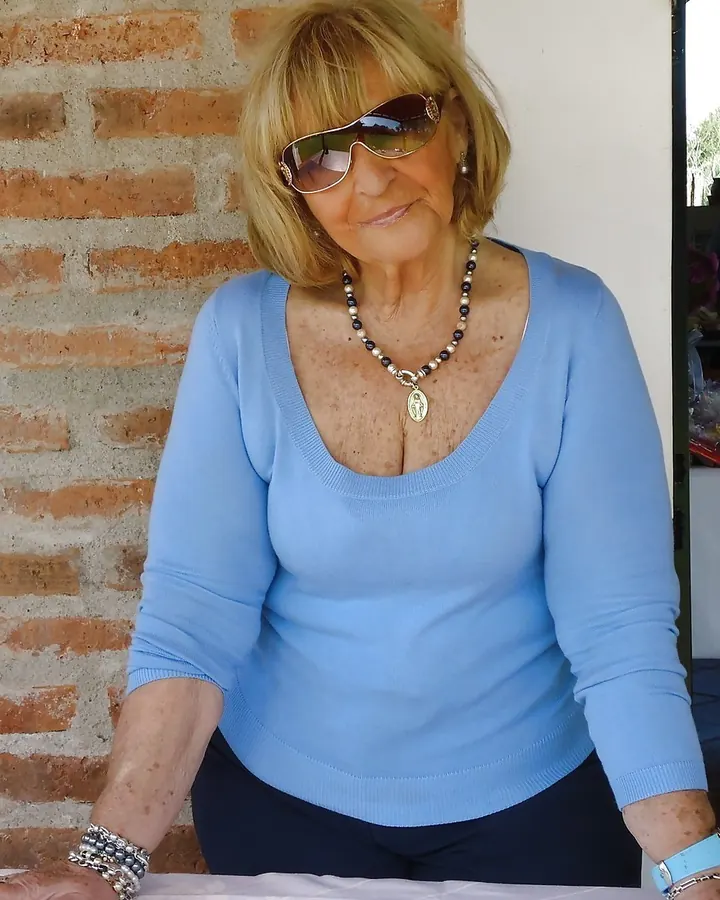 Femme aux cheveux blonds, portant un haut bleu, assise sur un banc de pierre en extérieur, regardant la caméra.