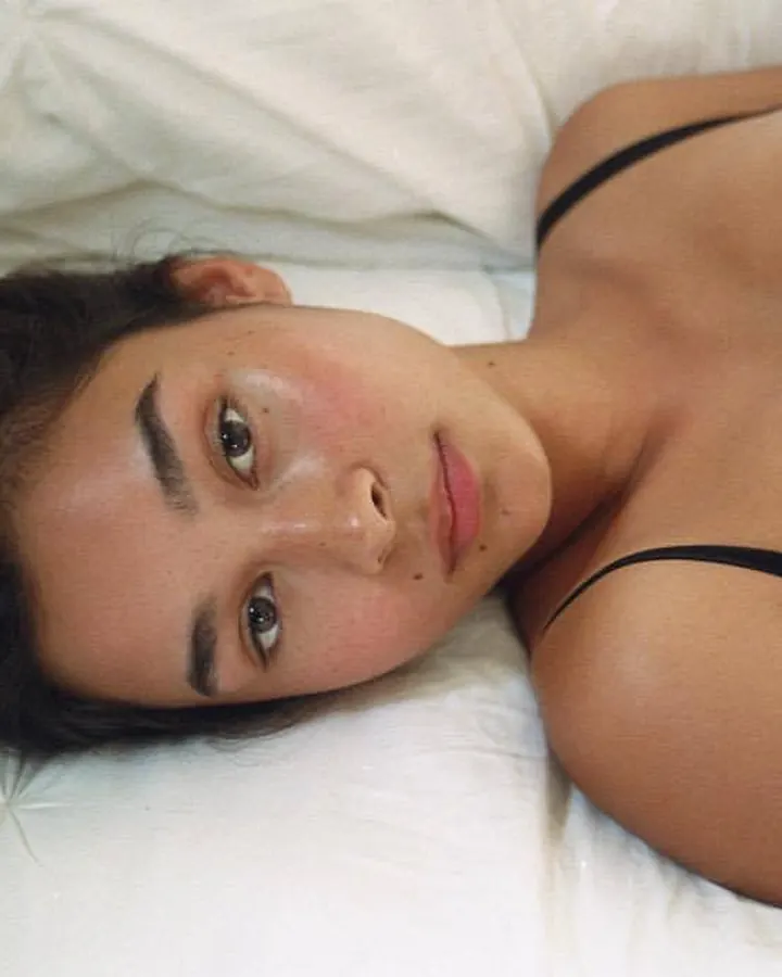 Femme aux cheveux bruns foncés, allongée sur un lit blanc, portant un haut noir, regardant directement l'objectif.