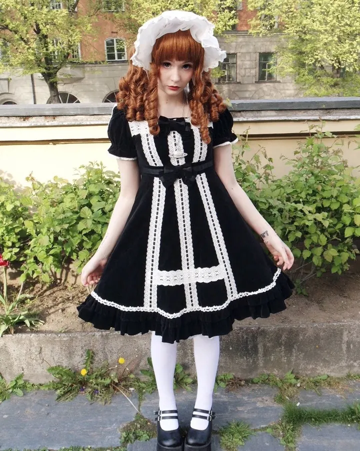 Femme aux cheveux roux, portant une robe noire et blanche avec un bonnet blanc, debout en extérieur, sur un trottoir pavé.