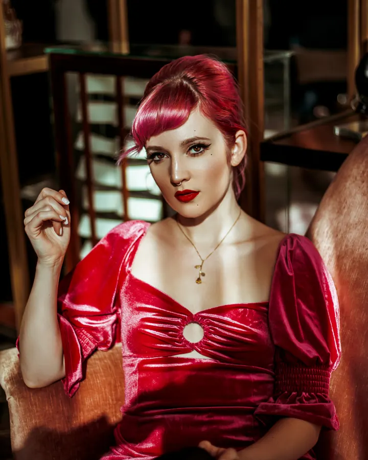 Femme aux cheveux roux, portant une robe rouge velours, assise sur un fauteuil, dans un décor sombre et chaleureux.