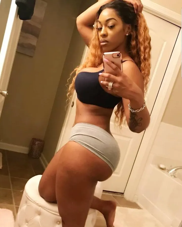 Femme aux cheveux longs ondulés, peau mate, portant un haut bleu marine et un short gris, prenant un selfie dans une salle de bain.