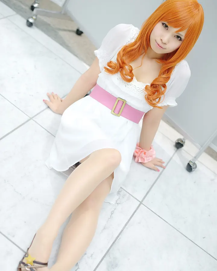 Femme asiatique aux cheveux roux, vêtue d'une robe blanche avec une ceinture rose, assise sur un sol carrelé blanc.