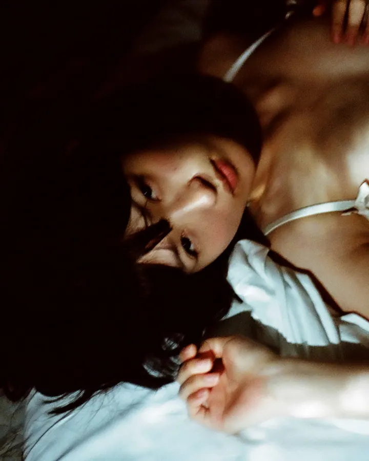 Femme asiatique aux cheveux bruns-noirs portant un haut noir et un jean, posant près d'une fenêtre avec des vitres texturées.