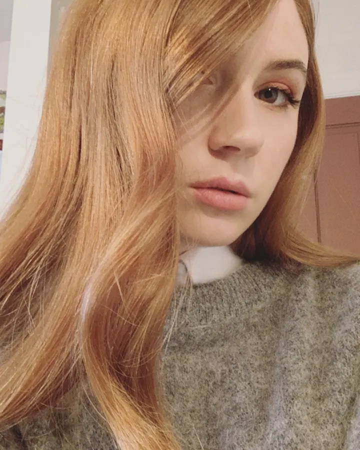 Femme européenne aux cheveux roux portant un haut noir et un jean, posant près d'une fenêtre avec des vitres texturées.