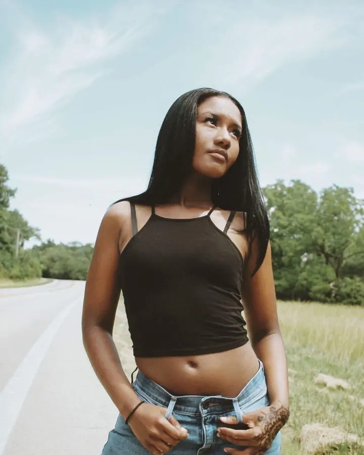 Femme asiatique aux cheveux bruns-noirs portant un haut noir et un jean, debout sur une route bordée d'arbres sous un ciel clair.