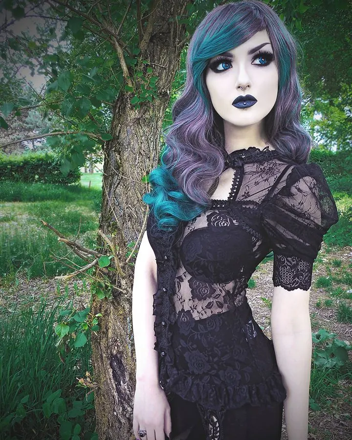 Femme aux cheveux roux portant une robe noire en dentelle, debout près d'un arbre dans un jardin verdoyant.