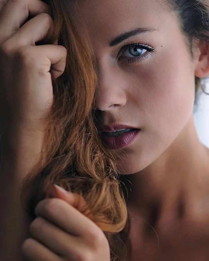 Femme aux yeux bleus et cheveux roux, nue, tenant ses cheveux avec une main, regardant directement l'appareil