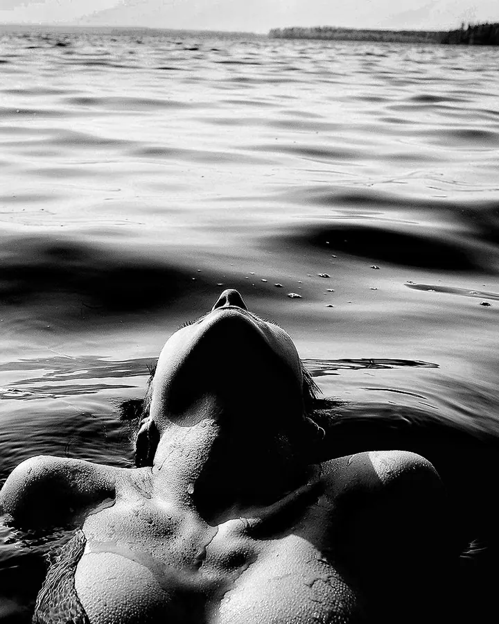 Femme aux cheveux bruns noirs, vue de face, flottant dans l’eau, le visage levé vers le ciel, photo en noir et blanc