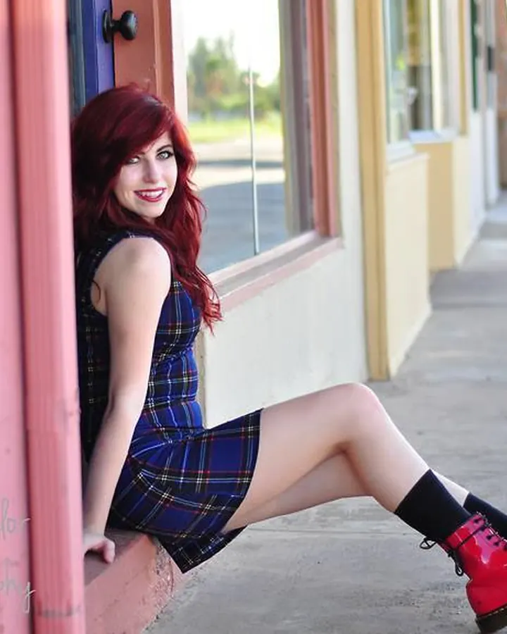 Jeune femme aux cheveux roux assise contre un mur coloré, vêtue d'une robe écossaise et de bottes rouges, souriante et provocante.
