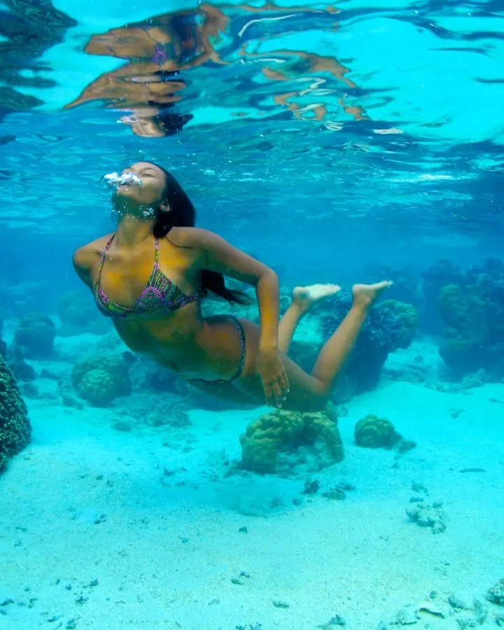 Femme en bikini coloré, plongeant sous l'eau turquoise, entourée de coraux, bulles autour de son visage.
