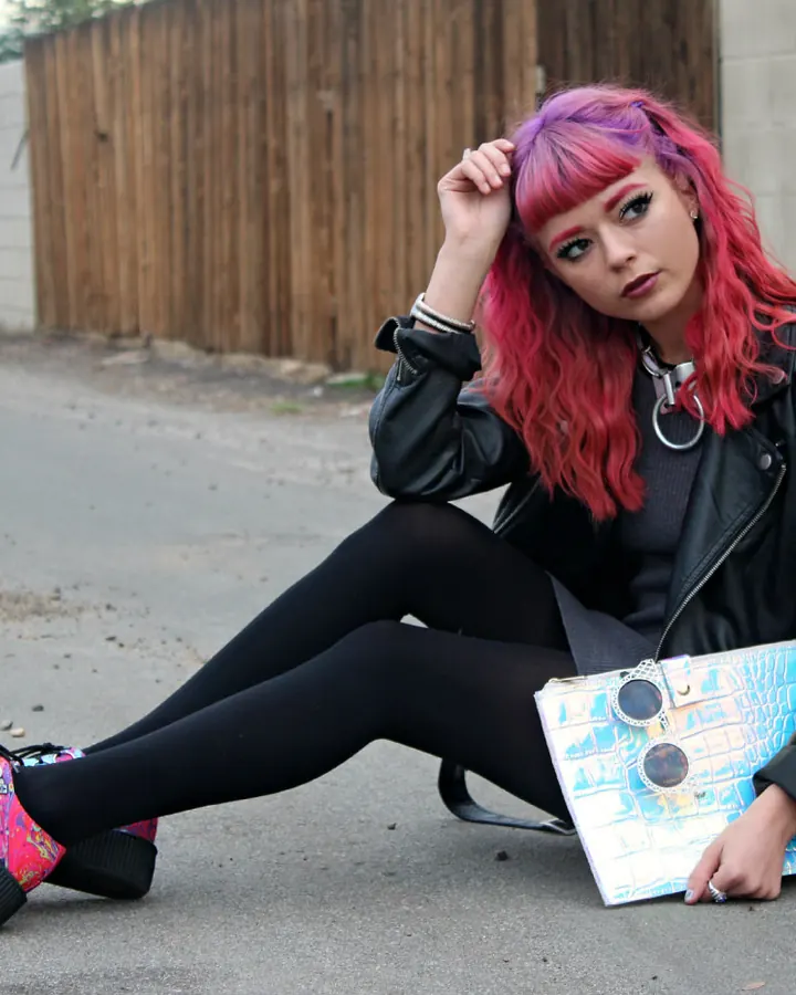 Femme aux cheveux roses et violets, vêtue d'une veste en cuir noir, pantalon noir, bottines colorées, assise sur un sol en béton, tenant un sac brillant.