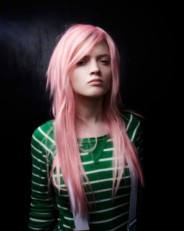 Femme aux cheveux roses, vêtue d'un haut rayé vert et blanc, regardant directement l'objectif avec un air neutre.