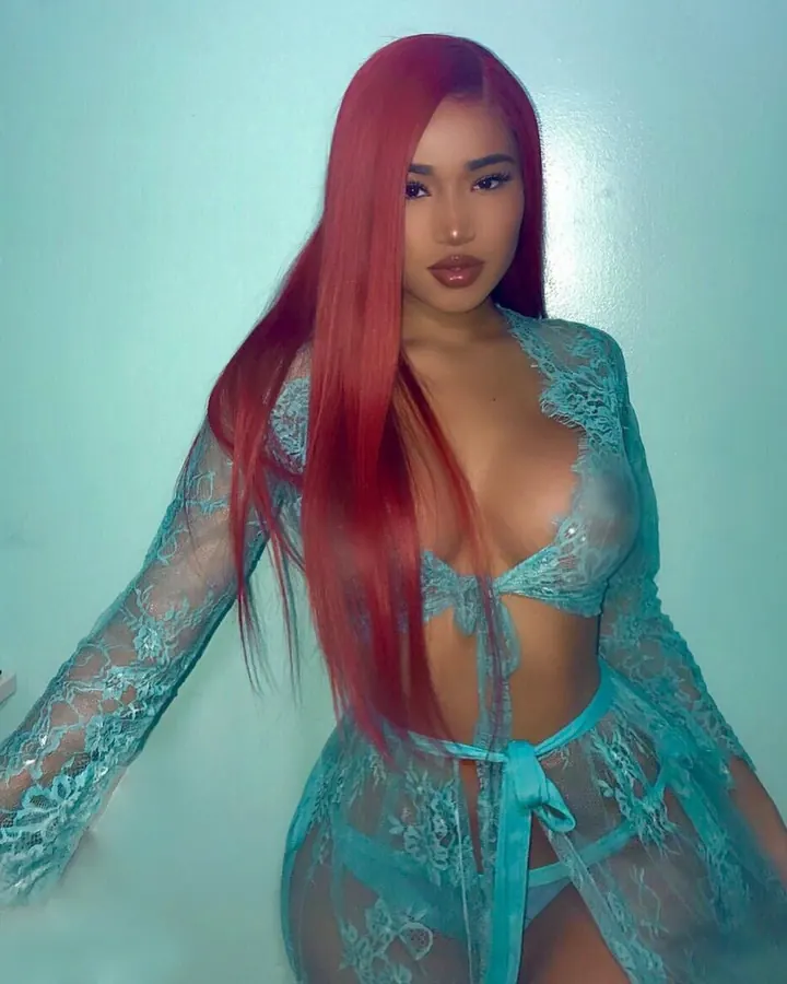 Femme aux cheveux rouges longs, vêtue d'une tenue turquoise en dentelle transparente, regardant directement l'objectif.