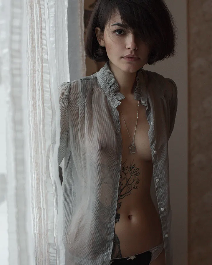 Femme aux cheveux bruns, portant une chemise transparente, posant près d'une fenêtre avec un tatouage visible sur le ventre.