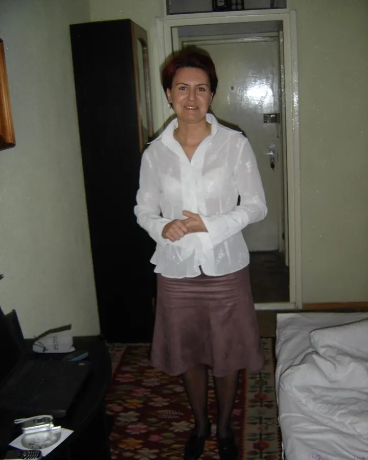 Femme aux cheveux roux, portant une chemise blanche transparente et une jupe marron, debout dans une chambre.