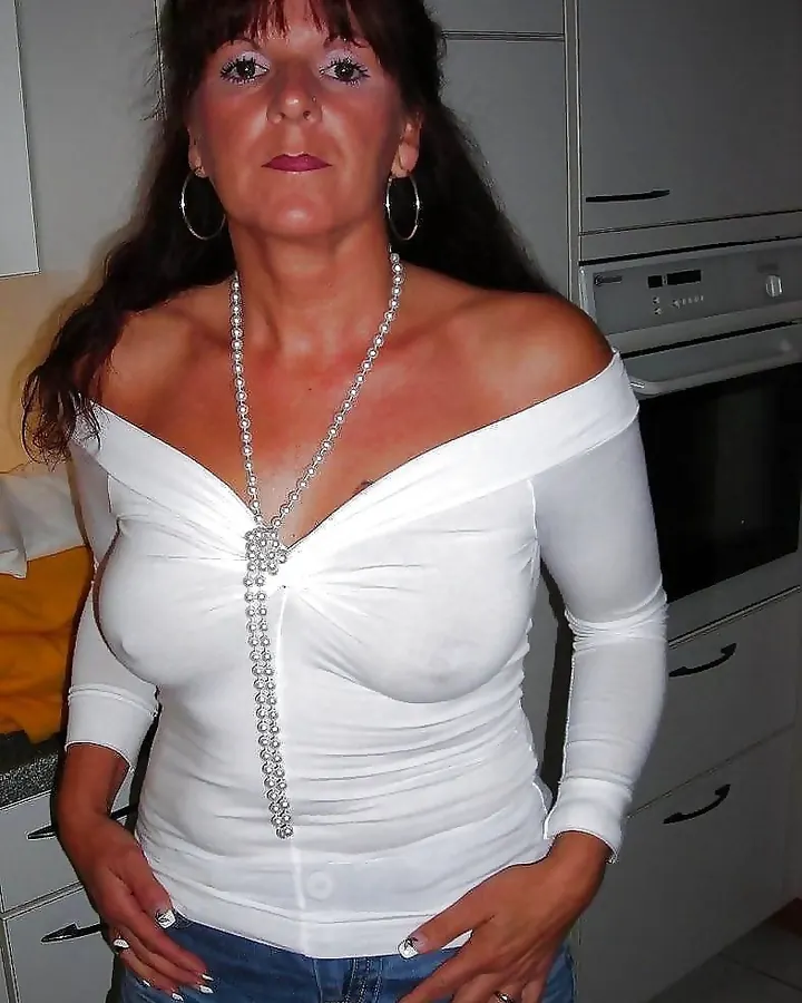 Femme aux cheveux bruns, portant un haut blanc décolleté et un jean, debout dans une cuisine.