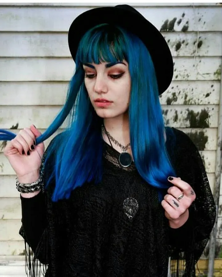 Femme aux cheveux bleus portant un chapeau noir, tenant une mèche de cheveux devant un mur en bois