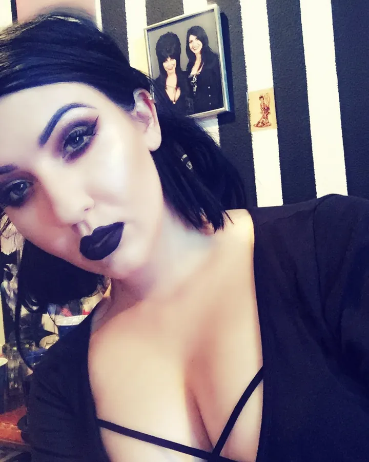 Femme aux cheveux noirs, maquillage sombre, portant un haut noir, prenant un selfie devant un mur rayé noir et blanc
